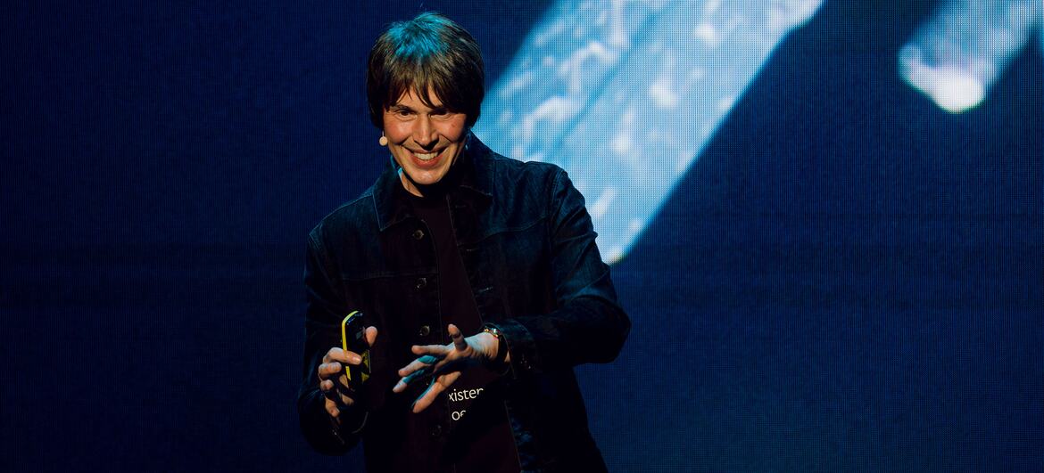 Professor Brian Cox, an English physicist and musician, smiling while giving a presentation on stage with a large screen behind him.