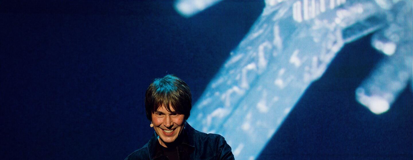 Professor Brian Cox, an English physicist and musician, smiling while giving a presentation on stage with a large screen behind him.