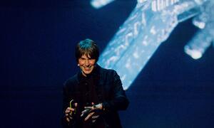Professor Brian Cox, an English physicist and musician, smiling while giving a presentation on stage with a large screen behind him.
