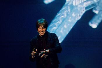 Professor Brian Cox, an English physicist and musician, smiling while giving a presentation on stage with a large screen behind him.