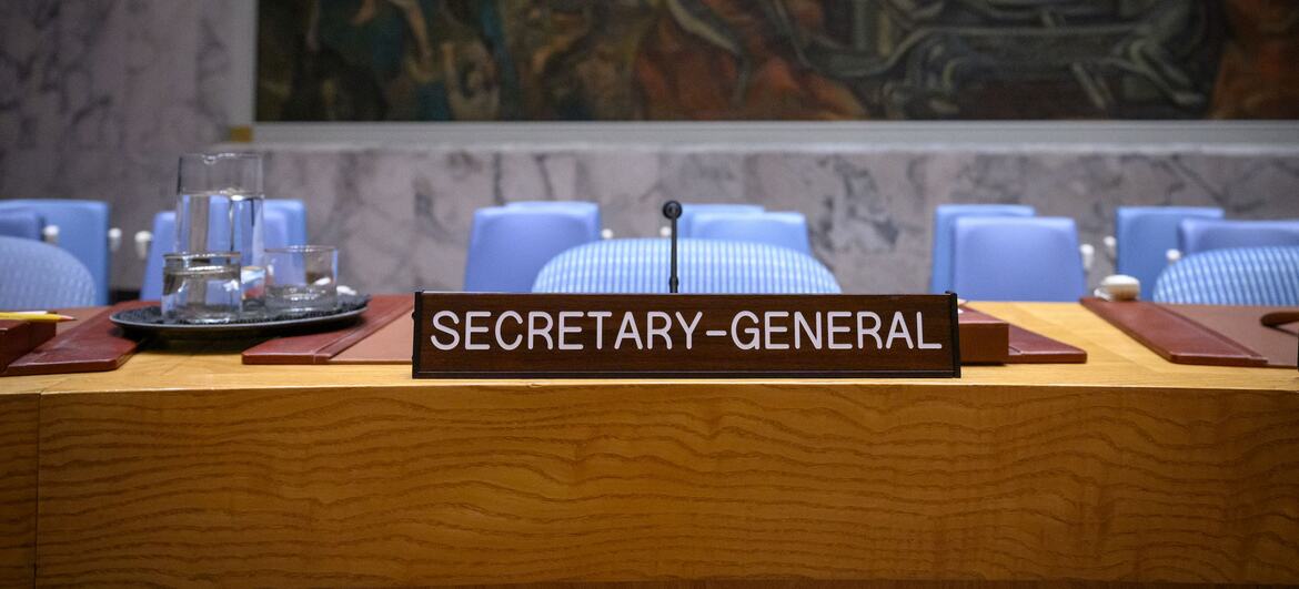 The Secretary-General's nameplate on a wooden desk in the UN Security Council Chamber, with blue chairs and a large mural in the background.