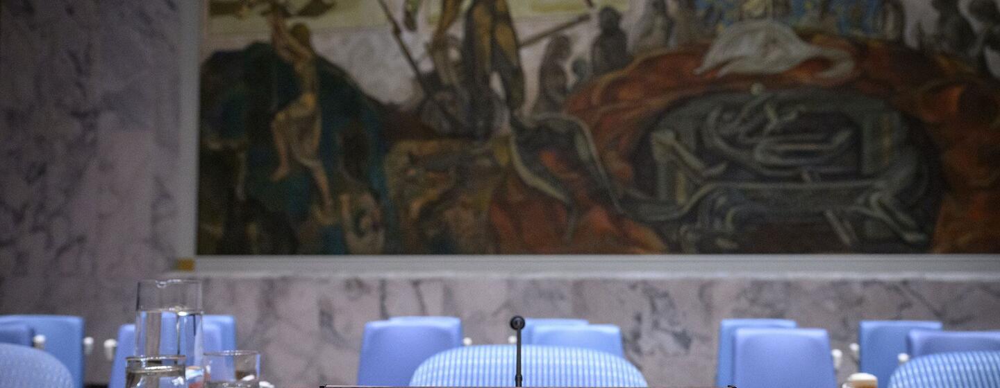 La plaque portant le nom du Secrétaire général sur un bureau en bois dans la salle du Conseil de sécurité de l'ONU, avec des chaises bleues et une grande fresque murale en arrière-plan.
