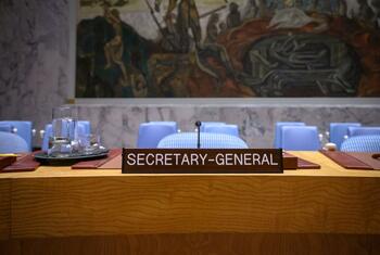 La placa con el nombre del Secretario General sobre un escritorio de madera en la Sala del Consejo de Seguridad de la ONU, con sillas azules y un gran mural en el fondo.