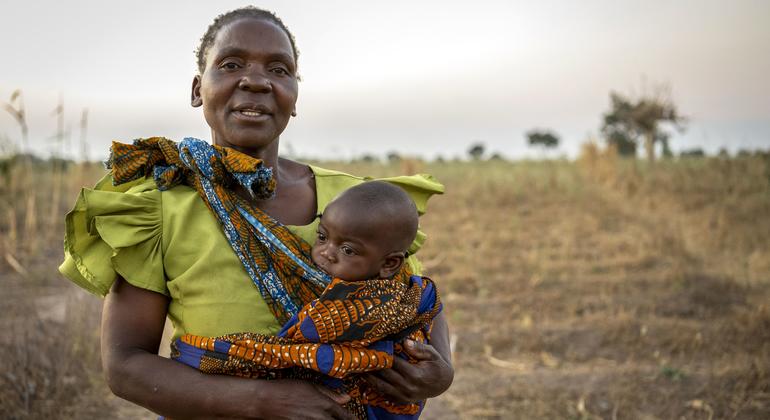 Mwanamke aliyevaa fulana ya kijani kibichi amembeba mtoto mchanga aliyefungwa kwa kitambaa chenye muundo, amesimama kwenye shamba kavu lenye miti michache nyuma.