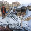 Un homme et une femme se trouvent dans un campement de fortune à Gaza. La femme trie un tas de couvertures et d'autres provisions, tandis que l'homme se tient à proximité. À l'arrière-plan, on aperçoit d'autres tentes et des bâtiments endommagés.