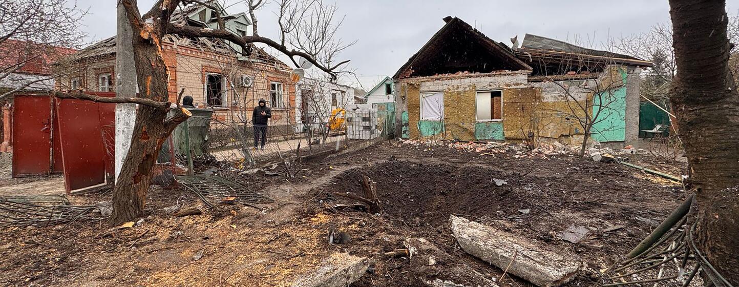 Un bâtiment résidentiel endommagé en Ukraine, avec un toit effondré et un grand cratère au premier plan, et une personne debout à l'arrière-plan.