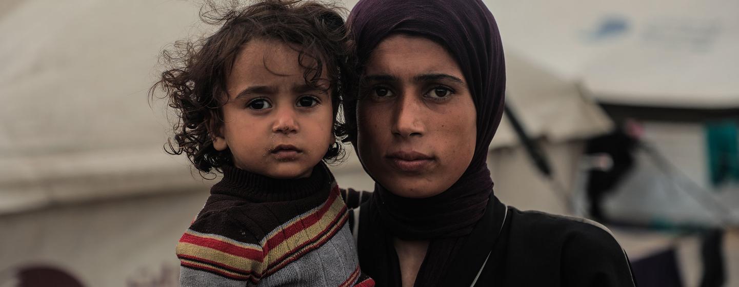 Une mère et son jeune enfant se tiennent debout dans un campement de fortune à Gaza. La mère tient l'enfant dans ses bras et tous deux regardent l'objectif avec gravité, une tente étant visible en arrière-plan.