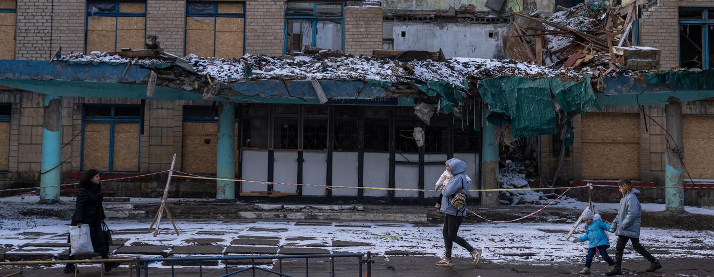 Le 1er janvier 2024, à Kurakhove, en Ukraine, une femme portant un enfant passe devant un bâtiment fortement endommagé. D'autres familles, dont des enfants, marchent dans la rue devant la structure en ruine.