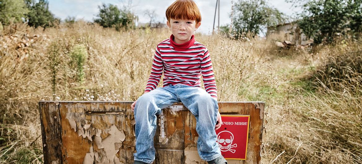 A young boy  in Kharkiv region, Ukraine, sits in front of a sign that reads "Danger of Mines."