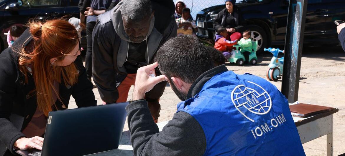 Um trabalhador humanitário da OIM com colete azul ajuda um homem e uma mulher com um laptop num balcão de matrícula ao ar livre, com outros deslocados e veículos no fundo.