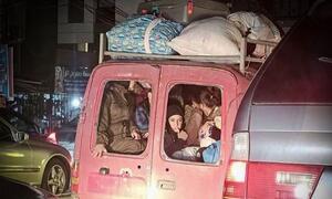 Un grupo de personas desplazadas, incluyendo mujeres y niños, se amontonan en la parte trasera de una furgoneta roja cargada de equipaje en el techo, ilustrando la crisis humanitaria en el Líbano.