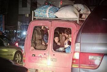 Un groupe de personnes déplacées, y compris des femmes et des enfants, sont entassées à l'arrière d'une camionnette rouge chargée de bagages sur le toit, illustrant la crise humanitaire au Liban.