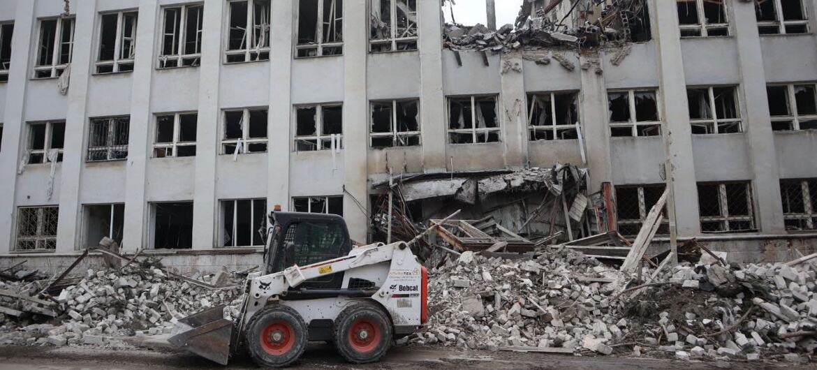 A white Bobcat skid steer loader is positioned in front of a large, multi-story building in Sloviansk, Ukraine, that has been heavily damaged with collapsed sections and shattered windows. Piles of concrete rubble and debris are scattered on the ground.