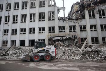 A white Bobcat skid steer loader is positioned in front of a large, multi-story building in Sloviansk, Ukraine, that has been heavily damaged with collapsed sections and shattered windows. Piles of concrete rubble and debris are scattered on the ground.