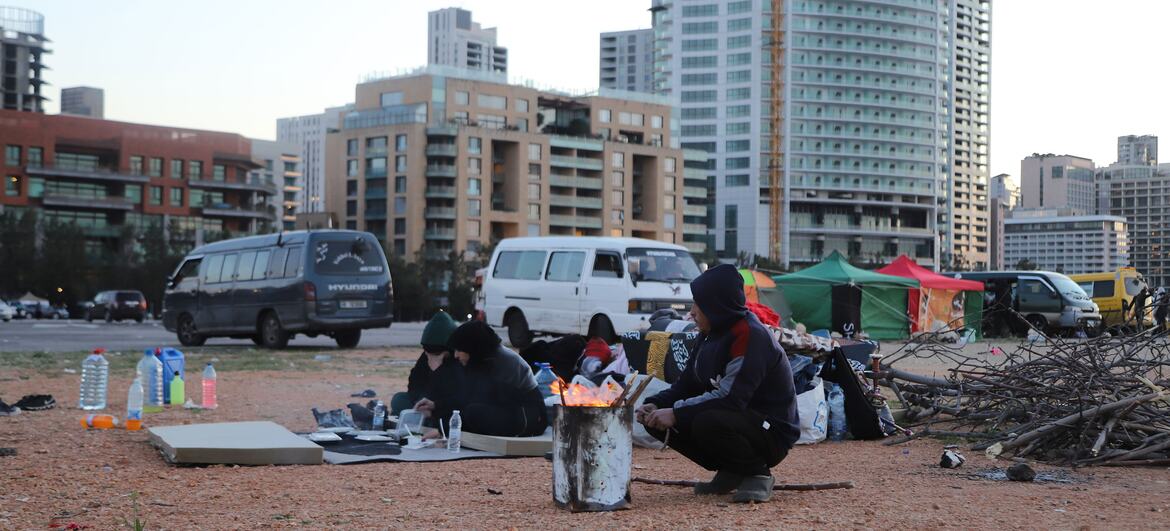 Displaced families in makeshift tents and shelters on a dusty lot in Beirut, Lebanon, following air strikes in the Dahiyeh suburb on March 9, 2026. People are seen cooking over a small fire with high-rise buildings in the background.