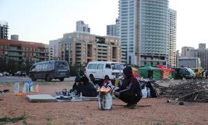 Displaced families in makeshift tents and shelters on a dusty lot in Beirut, Lebanon, following air strikes in the Dahiyeh suburb on March 9, 2026. People are seen cooking over a small fire with high-rise buildings in the background.