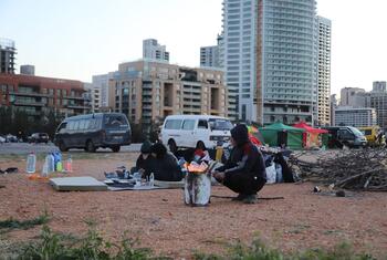 Displaced families in makeshift tents and shelters on a dusty lot in Beirut, Lebanon, following air strikes in the Dahiyeh suburb on March 9, 2026. People are seen cooking over a small fire with high-rise buildings in the background.