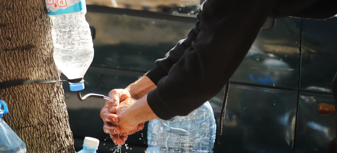 A man washes his hands using a makeshift water dispenser created from a plastic bottle attached to a tree, with several other large water bottles nearby. This scene depicts a humanitarian aid effort for displaced families in Beirut, Lebanon.