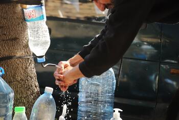 A man washes his hands using a makeshift water dispenser created from a plastic bottle attached to a tree, with several other large water bottles nearby. This scene depicts a humanitarian aid effort for displaced families in Beirut, Lebanon.