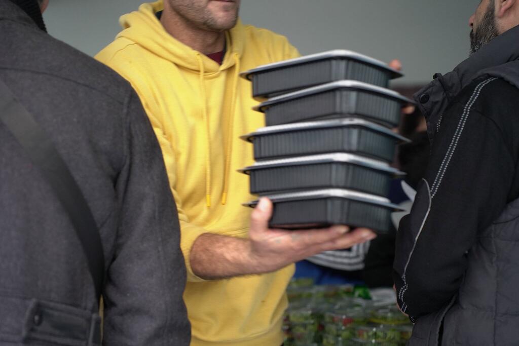 Un homme en hoodie jaune porte une pile de cinq conteneurs noirs de nourriture pendant une distribution de nourriture du PAM aux familles déplacées qui se réfugient dans une école publique de Tariq Jdide, Beyrouth, au Liban, le 7 mars 2026.