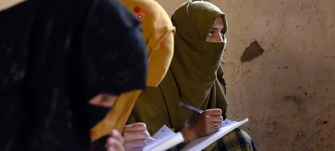 Des femmes et des filles afghanes portant des hijabs et des niqabs sont assises sur le sol en train d'écrire des notes dans une salle de classe.