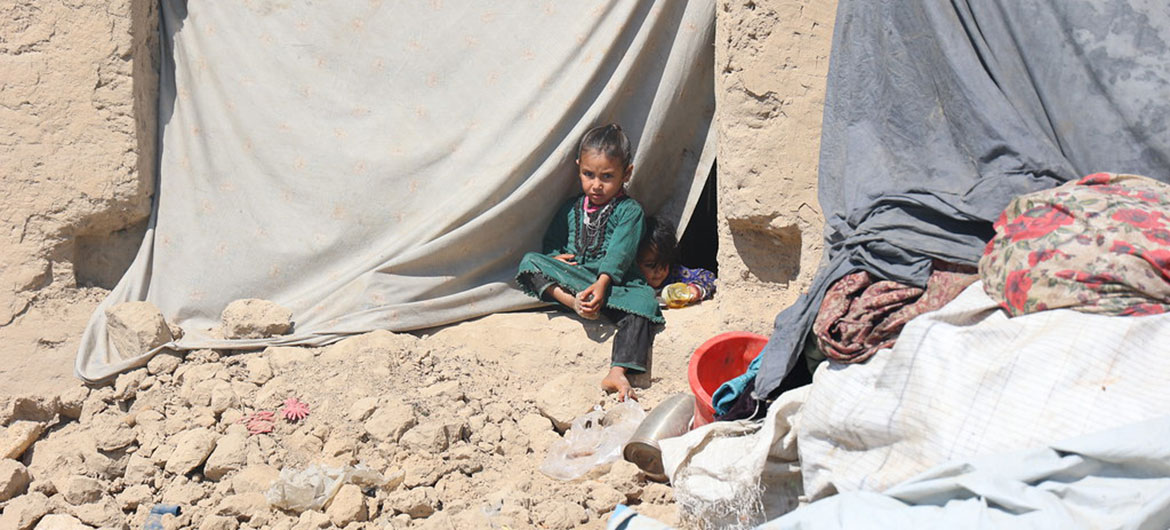 Two young girls sit at the entrance of a makeshift shelter made of mud brick and draped fabric, surrounded by rubble, a red basin, and blankets in Afghanistan.