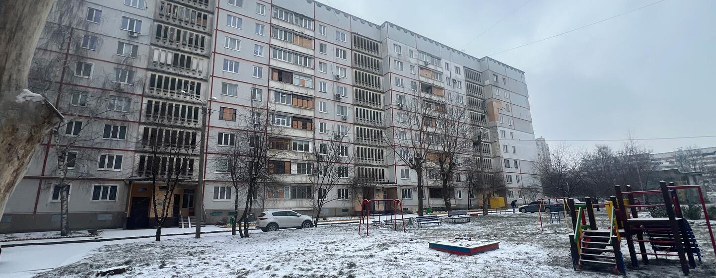 Edifícios de apartamentos danificados em Saltivka, Kharkiv, Ucrânia, com terreno coberto de neve e um parque infantil no primeiro plano.