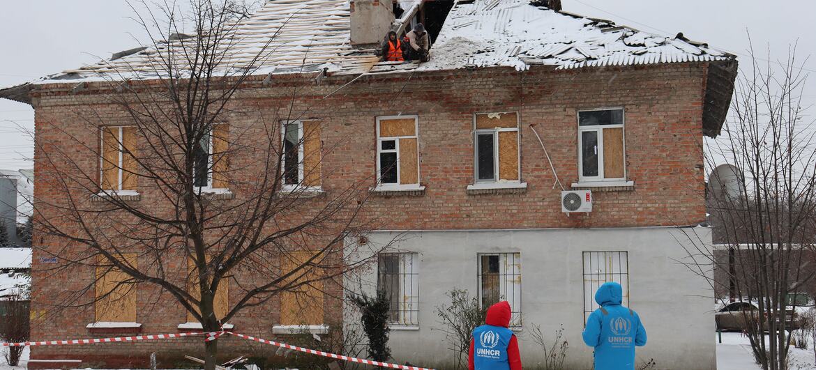 Two UNHCR workers in blue jackets stand near a damaged residential building in Dnipro, Ukraine, covered in snow. The roof is partially collapsed and windows are boarded up, with workers inspecting the damage.