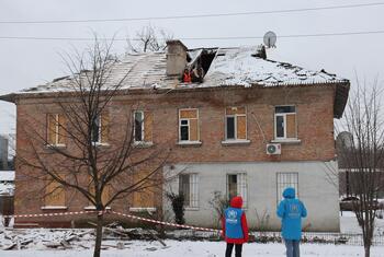 Dos trabajadores del ACNUR con chaquetas azules se encuentran cerca de un edificio residencial dañado en Dnipro, Ucrania, cubierto de nieve.