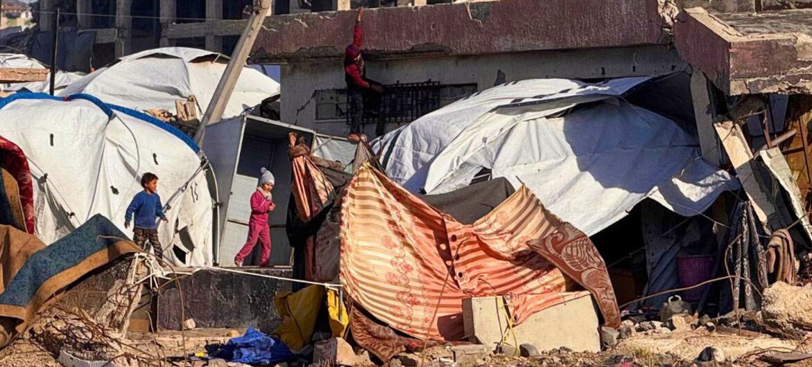 Crianças caminham perto de tendas improvisadas construídas entre os escombros e edifícios destruídos ao longo da praia de Gaza, destacando as duras condições de vida dos palestinos deslocados.