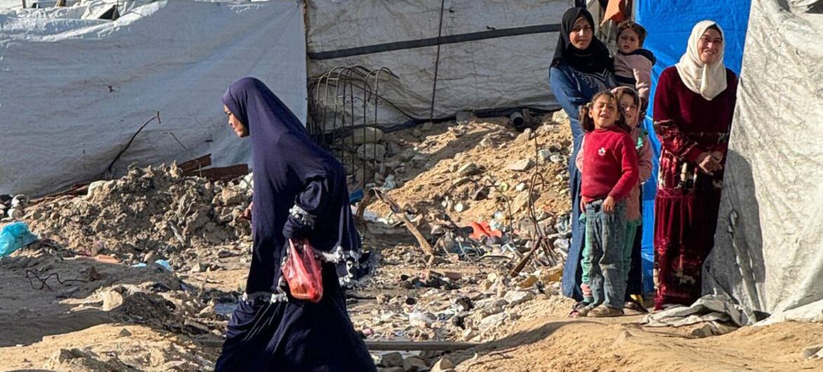 A woman walks through a muddy, debris-strewn area near makeshift tents where displaced Palestinians are living in harsh conditions in Gaza.