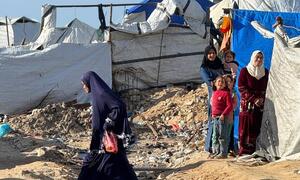 A woman walks through a muddy, debris-strewn area near makeshift tents where displaced Palestinians are living in harsh conditions in Gaza.