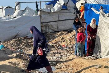 A woman walks through a muddy, debris-strewn area near makeshift tents where displaced Palestinians are living in harsh conditions in Gaza.
