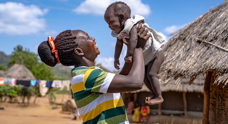 Радостная мать в Уганде, держащая своего ребенка высоко, улыбаясь, символизируя расширение прав и возможностей через программу NutriCash, которая поддерживает беременных и кормящих матерей денежными переводами и ресурсами для улучшения питания и создания малого бизнеса.
