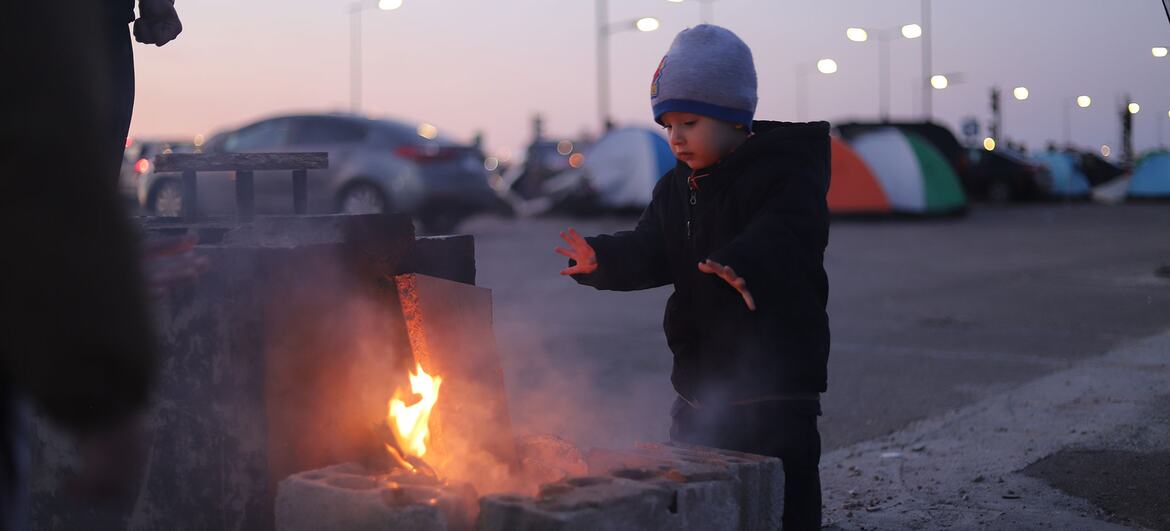 A young child warms their hands over a small fire built from concrete blocks in a parking lot, with tents and other displaced people visible in the background in Beirut, Lebanon on March 9, 2026.