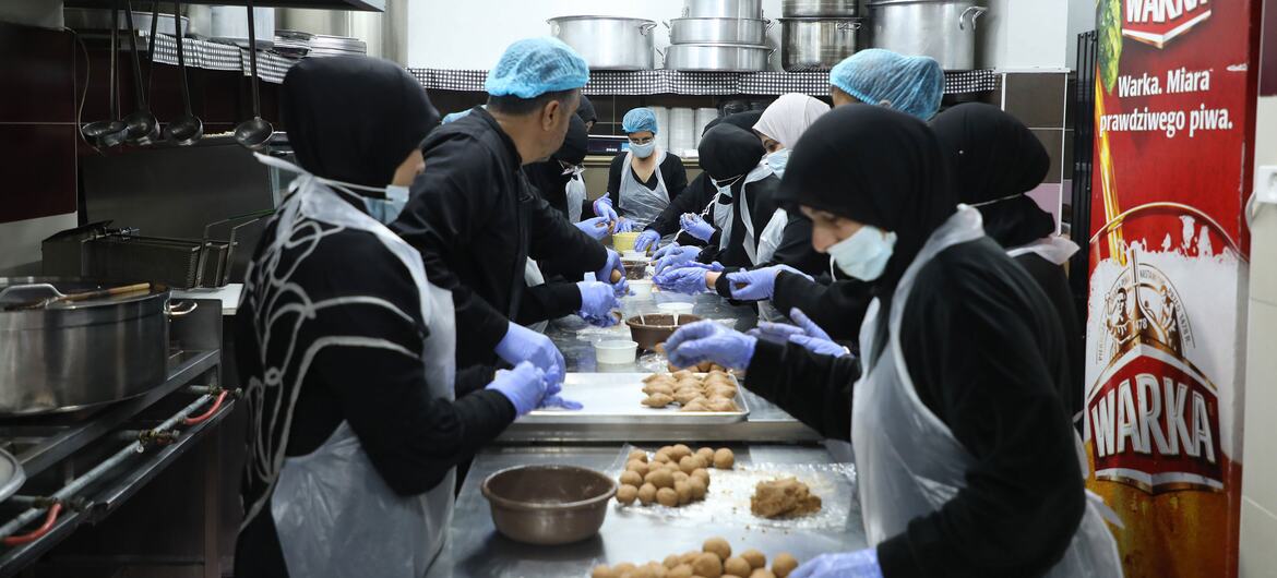 Mujeres con hijab, mascarillas y guantes preparan comida en una gran cocina comercial.