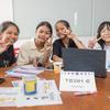 Quatro jovens estudantes em um evento escolar no Camboja, sorrindo e fazendo o sinal de paz para a câmera. Elas estão sentadas ao redor de uma mesa com laptops, papéis e uma placa que diz "EQUIPE 6" para uma competição de design de videogame.