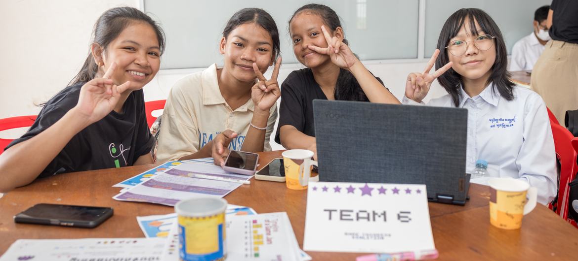 Quatro jovens estudantes em um evento escolar no Camboja, sorrindo e fazendo o sinal de paz para a câmera. Elas estão sentadas ao redor de uma mesa com laptops, papéis e uma placa que diz "EQUIPE 6" para uma competição de design de videogame.
