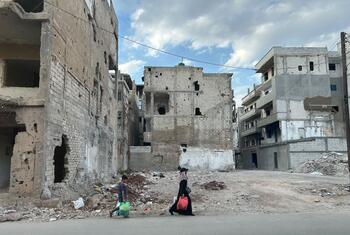 Two individuals walk through a street in Homs, Syria, past heavily damaged and destroyed buildings, highlighting the aftermath of conflict.