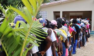 Una larga fila de personas espera al aire libre en Haití para recibir dinero en efectivo como parte del programa "dinero por trabajo" del Programa Mundial de Alimentos (PMA), con plantas tropicales verdes en primer plano.