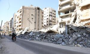 A street in Dahiyeh, Beirut, Lebanon, showing massive destruction and rubble from air strikes. A person rides a motorcycle past the collapsed buildings and debris.