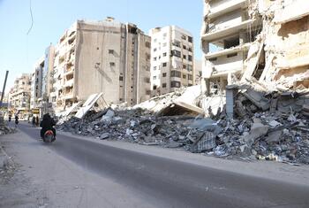 A street in Dahiyeh, Beirut, Lebanon, showing massive destruction and rubble from air strikes. A person rides a motorcycle past the collapsed buildings and debris.