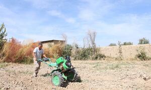 A man operating a green motorized tiller in a dry, tilled field under a blue sky.