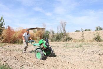 A man operating a green motorized tiller in a dry, tilled field under a blue sky.