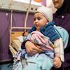 طفل يبلغ من العمر 10 أشهر، وذراعُه ملفوفة بالجبس، يُحمله والدته في غرفة بمستشفى. الطفل ملفوف في بطانيات ويرتدي قبعة صوفية.