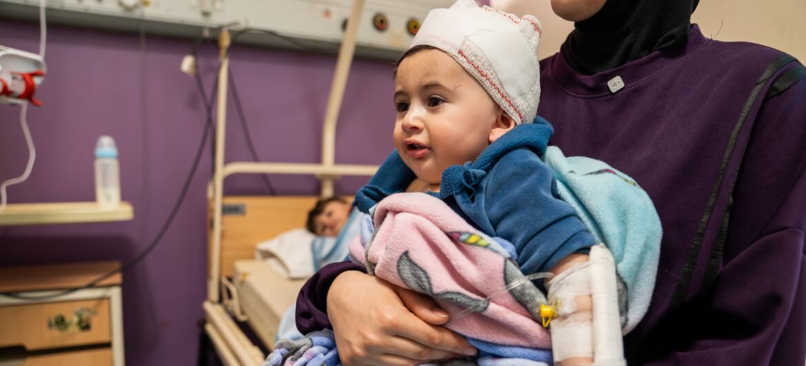 طفل يبلغ من العمر 10 أشهر، وذراعُه ملفوفة بالجبس، يُحمله والدته في غرفة بمستشفى. الطفل ملفوف في بطانيات ويرتدي قبعة صوفية.