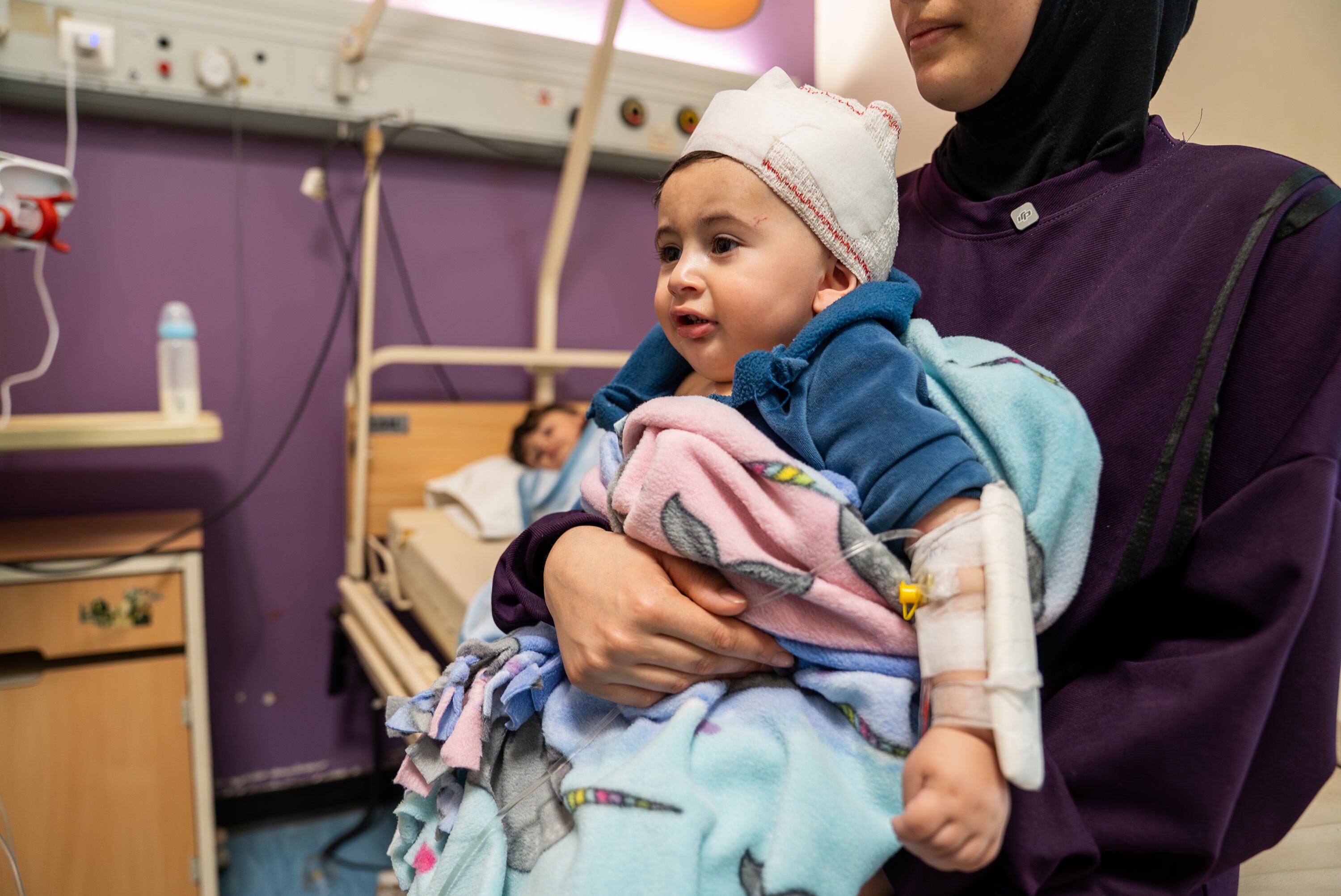 A 10-month-old baby, arm in a cast, is carried by his mother in a hospital room. The child is wrapped in blankets and wearing a wool hat.
