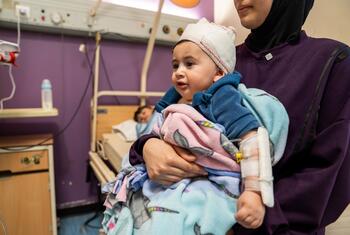 طفل يبلغ من العمر 10 أشهر، وذراعُه ملفوفة بالجبس، يُحمله والدته في غرفة بمستشفى. الطفل ملفوف في بطانيات ويرتدي قبعة صوفية.