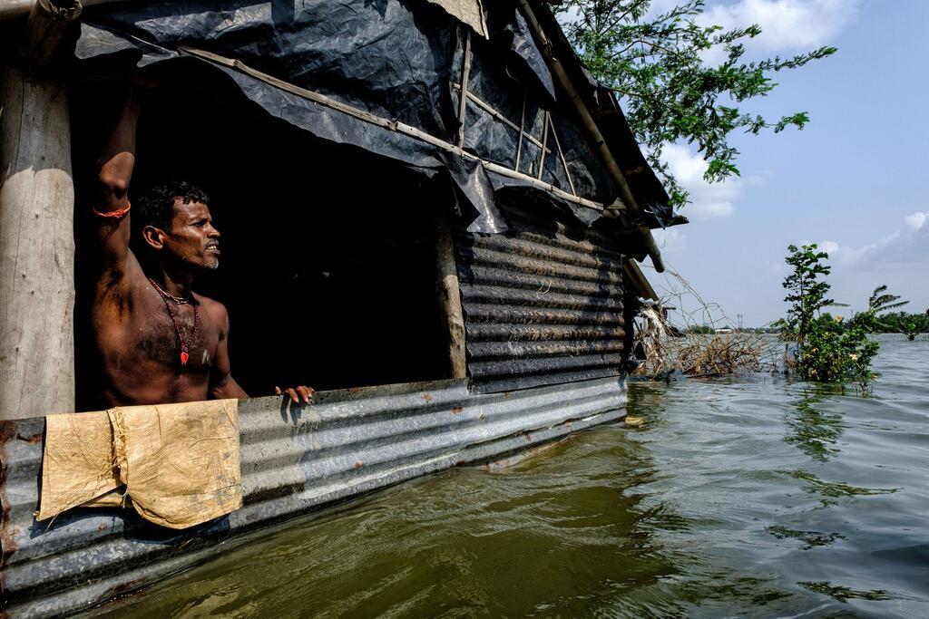 A shirtless man stands in the doorway of his flooded tin-roofed home in a coastal village in the Sundarbans, India, surrounded by floodwaters from sea level rise and cyclones.