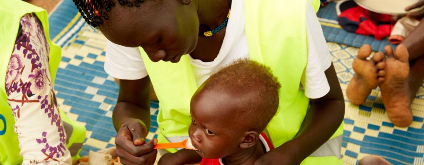 Un travailleur humanitaire portant un gilet jaune nourrit un enfant malnutri à la cuillère dans le cadre d'un programme de nutrition au Soudan du Sud.
